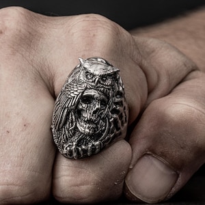 Può includere: Un anello d'argento con un designo dettagliato di un gufo appollaiato su un teschio. Il gufo ha un'espressione feroce e le sue ali sono spiegate.