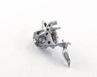 Op de afbeelding: Een zilveren bedel in de vorm van een tatoeagemachine. De bedel is gedetailleerd en realistisch, met een naald en een handvat.