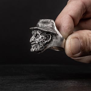 May include: A silver ring featuring a detailed, three-dimensional sculpture of a horror movie character's face wearing a hat.