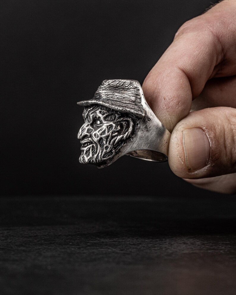 May include: A silver ring featuring a detailed, three-dimensional sculpture of a horror movie character's face wearing a hat.