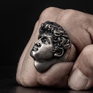 May include: A silver ring featuring a detailed sculpture of a man's face. The ring is worn on a person's finger.