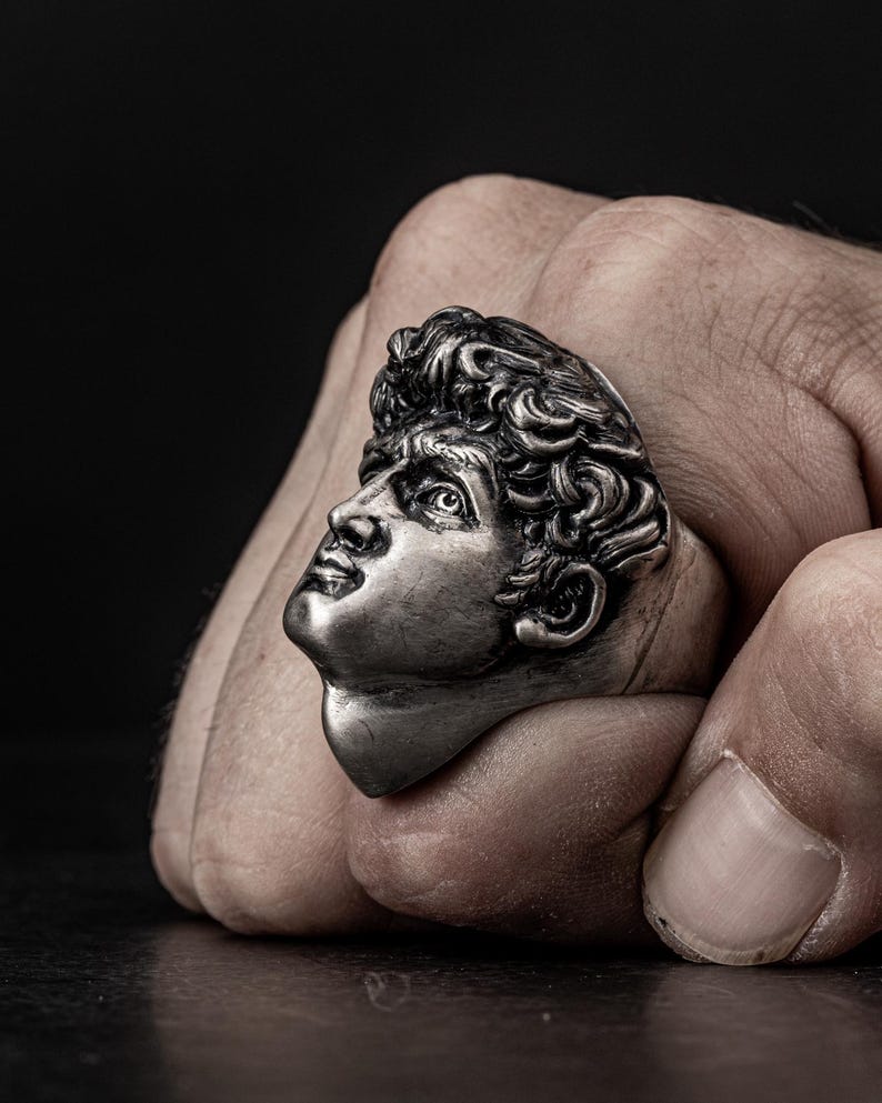 May include: A silver ring featuring a detailed sculpture of a man's face. The ring is worn on a person's finger.