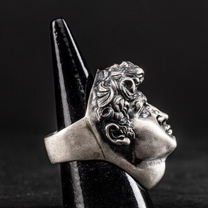 May include: A silver ring with a detailed sculpture of a man's face. The ring is on a black ring display stand.