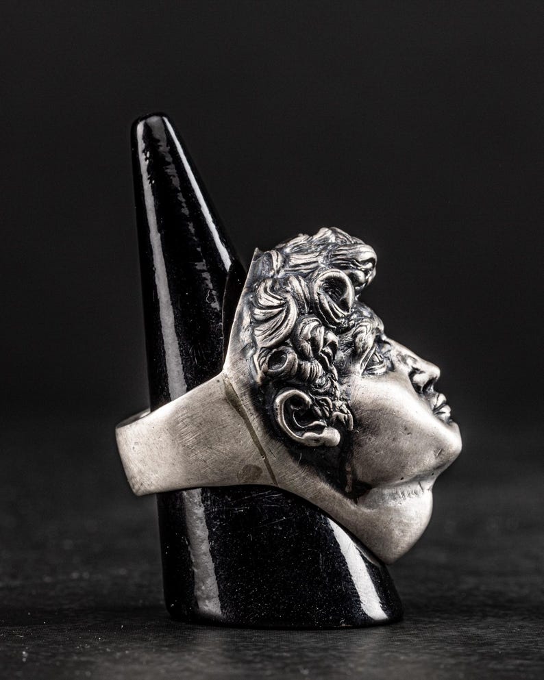 May include: A silver ring with a detailed sculpture of a man's face. The ring is on a black ring display stand.