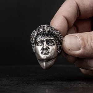 May include: A silver-colored sculpture of a man's head with short, curly hair. The sculpture is detailed and realistic, with a serious expression on the man's face.