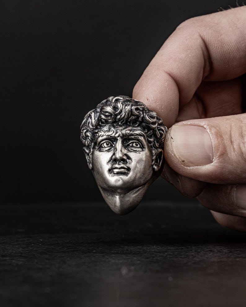 May include: A silver-colored sculpture of a man's head with short, curly hair. The sculpture is detailed and realistic, with a serious expression on the man's face.