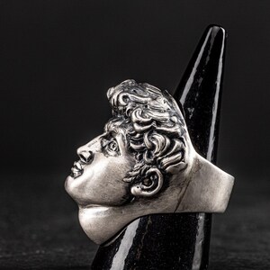 May include: A silver ring featuring a detailed sculpture of a man's face. The ring is displayed on a black cone-shaped stand.
