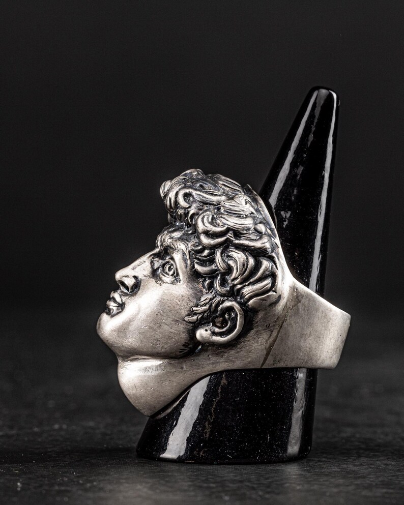 May include: A silver ring featuring a detailed sculpture of a man's face. The ring is displayed on a black cone-shaped stand.