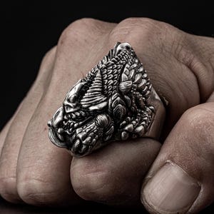May include: A silver ring with a detailed dragon design. The dragon's head is facing forward with its mouth open and its wings spread out.