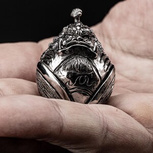 May include: A silver-coloured, detailed, ornate, decorative figurine of a mythical creature with a fierce expression. The figurine is shaped like an egg and has a textured surface.