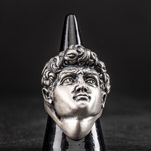 May include: A silver ring featuring a detailed sculpture of a man's face. The ring is displayed on a black cone-shaped stand.
