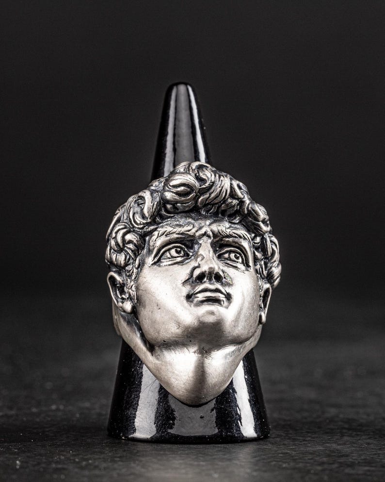 May include: A silver ring featuring a detailed sculpture of a man's face. The ring is displayed on a black cone-shaped stand.
