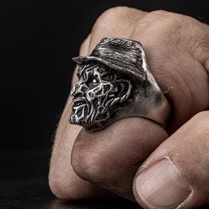 May include: A silver ring featuring a detailed, three-dimensional sculpture of a horror movie character's head wearing a hat.