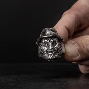 May include: A silver ring with a detailed, sculpted face of a horror movie character wearing a hat. The character has a menacing expression with sharp teeth and dark eyes.