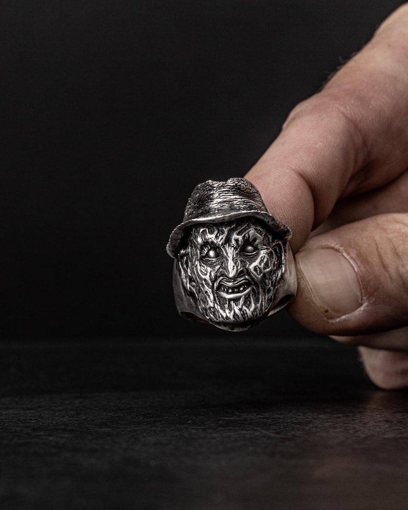 May include: A silver ring with a detailed, sculpted face of a horror movie character wearing a hat. The character has a menacing expression with sharp teeth and dark eyes.