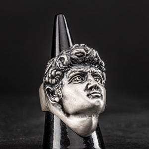 May include: A silver ring featuring a detailed sculpture of a man's face. The ring is displayed on a black cone-shaped stand.