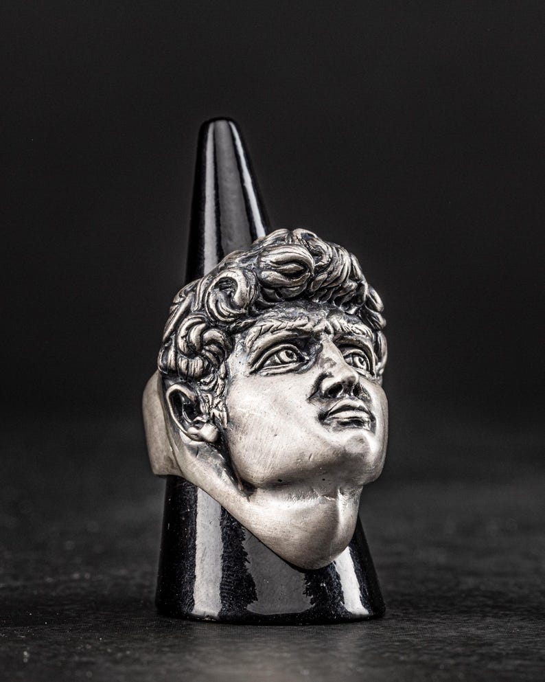 May include: A silver ring featuring a detailed sculpture of a man's face. The ring is displayed on a black cone-shaped stand.