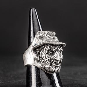 May include: A silver ring featuring a detailed, textured, and slightly menacing face with a hat. The ring is on a black display stand.
