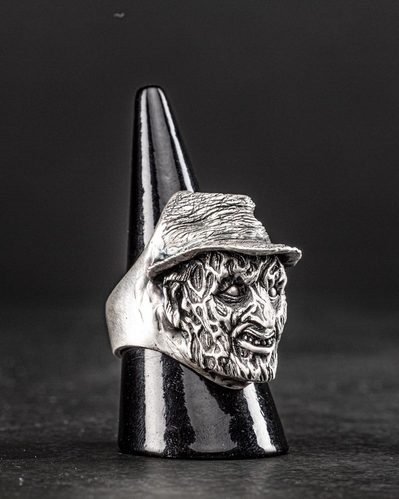 May include: A silver ring featuring a detailed, textured, and slightly menacing face with a hat. The ring is on a black display stand.