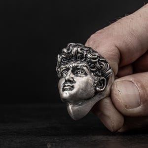 May include: A silver ring featuring a detailed sculpture of a man's face. The ring is worn on a finger and the face is looking up.