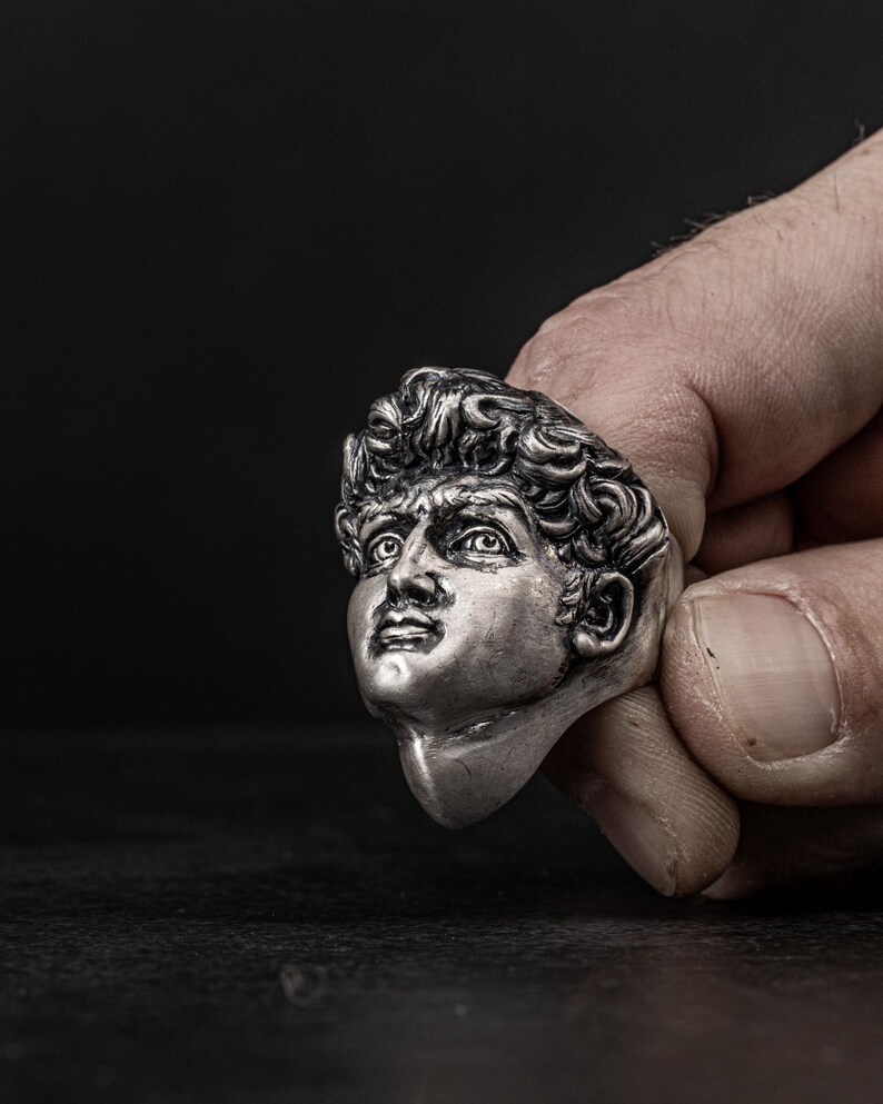 May include: A silver ring featuring a detailed sculpture of a man's face. The ring is worn on a finger and the face is looking up.