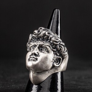 May include: A silver ring featuring a detailed sculpture of a man's face. The ring is displayed on a black cone-shaped stand.
