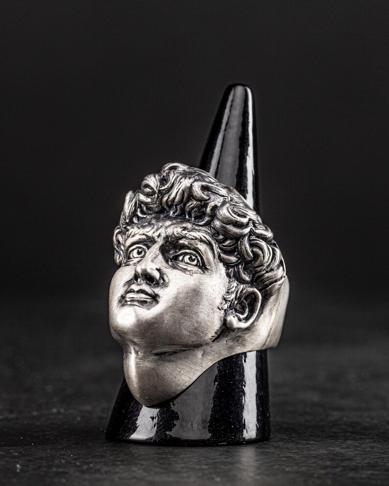 May include: A silver ring featuring a detailed sculpture of a man's face. The ring is displayed on a black cone-shaped stand.