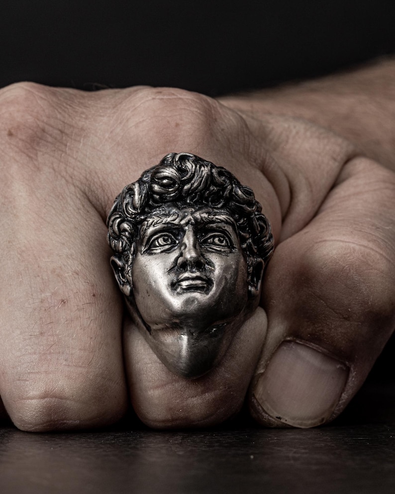 May include: A silver ring featuring a detailed sculpture of a man's face. The ring is worn on a person's finger.