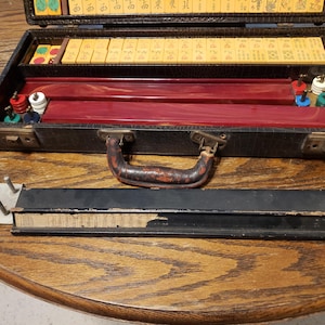 May include: An open vintage Mahjong set in a black case with a brown handle. The case contains tiles, red and brown game pieces, and a black and silver rack. The tiles are yellow with black characters. The set is on a wooden table.