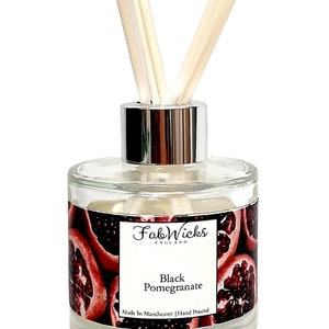 Puede incluir: Una botella de difusor de vidrio transparente con tapa plateada y palitos negros. La botella tiene una etiqueta que dice "Fab Wicks England" y "Black Pomegranate". La etiqueta también dice "Made in Manchester | Hand Poured". La botella está rodeada de un patrón de granadas rojas.