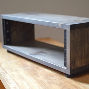 May include: A rectangular, open-front wooden shelf with a grey-stained top and sides. The shelf has dark brown sides and is supported by small black feet. Metal brackets are visible on the interior sides. The shelf is designed to hold audio equipment.