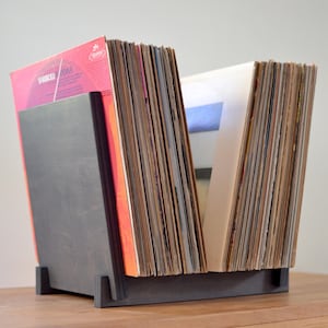 May include: A dark gray wooden vinyl record storage stand holding two rows of records. The stand features a solid base and two upright side panels that support the collection of albums, which include various colorful covers and white inner sleeves.