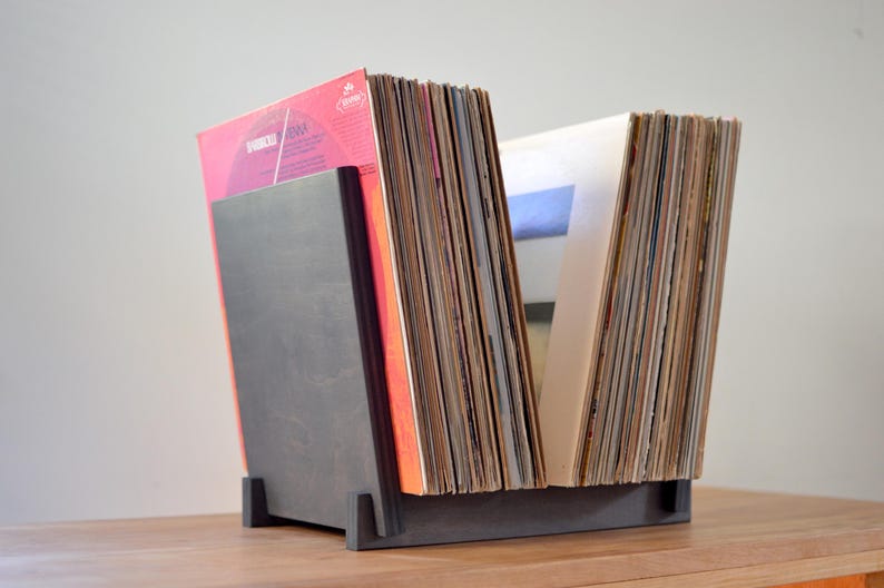May include: A dark gray wooden vinyl record storage stand holding a collection of various music albums. The stand features a solid front panel and a base with two upright supports, keeping the records organized in an upright, angled position.