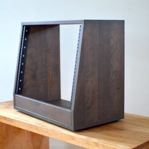 May include: A dark brown wooden rack unit with a rectangular frame and angled sides. The unit has metal strips with screw holes on the sides. It is sitting on a light brown wooden table.