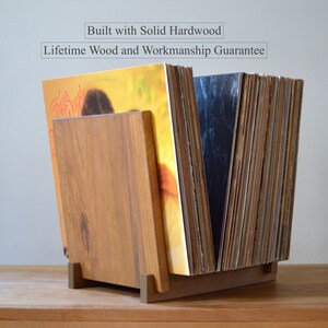 May include: A solid hardwood vinyl record storage stand holding a collection of records. The text above the stand reads: Built with Solid Hardwood, Lifetime Wood and Workmanship Guarantee. The stand features a natural wood grain finish.
