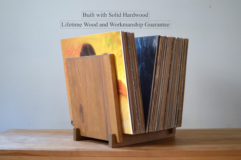 May include: A solid hardwood vinyl record storage stand holding a collection of records. The text above the stand reads: Built with Solid Hardwood, Lifetime Wood and Workmanship Guarantee. The stand features a natural wood grain finish.