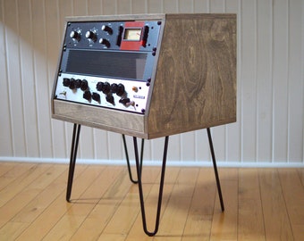 Puede incluir: Una consola de mezcla de audio de estilo vintage con botones negros y una pantalla roja y blanca, montada en un soporte de madera con patas de horquilla negras.