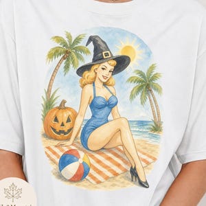 May include: White t-shirt featuring a watercolor illustration of a woman in a witch hat and blue swimsuit on a beach with a pumpkin, palm trees, and a beach ball. The design has a vintage, retro aesthetic.