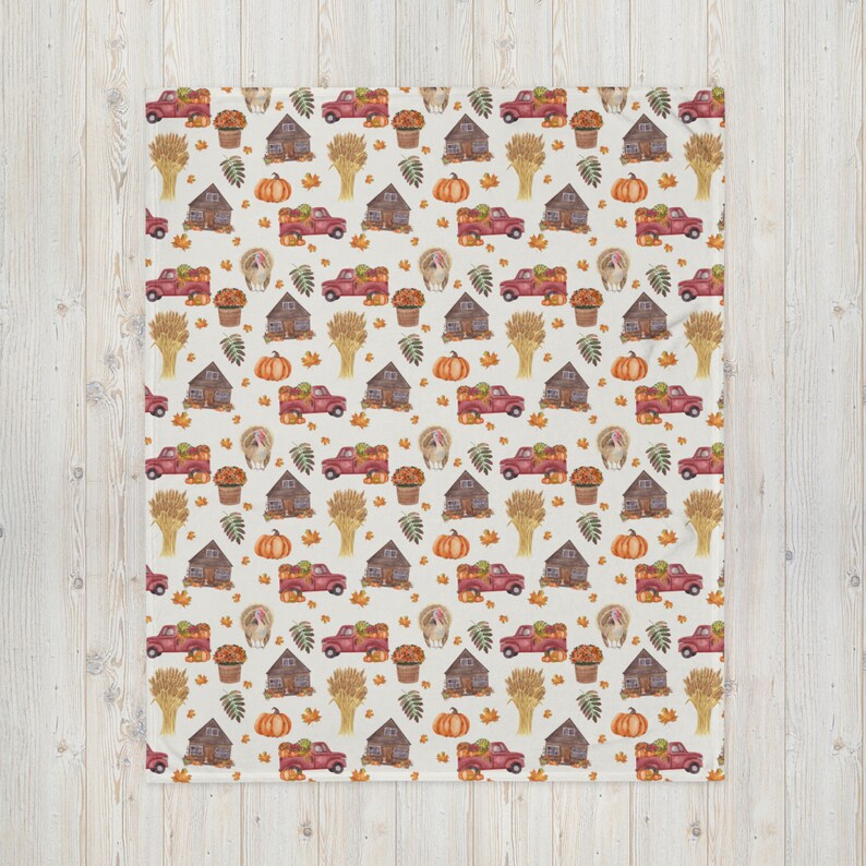 May include: A white throw blanket featuring a repeating watercolor pattern of red pickup trucks carrying pumpkins, brown rustic cabins, orange pumpkins, golden wheat sheaves, turkeys, potted orange flowers, and scattered autumn leaves.