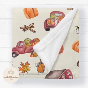 May include: A cream-colored throw blanket featuring a repeating watercolor pattern of red trucks carrying pumpkins, a corgi, cinnamon sticks, maple leaves, mugs of cider, and a red barn. The blanket has a white plush underside and a folded corner.