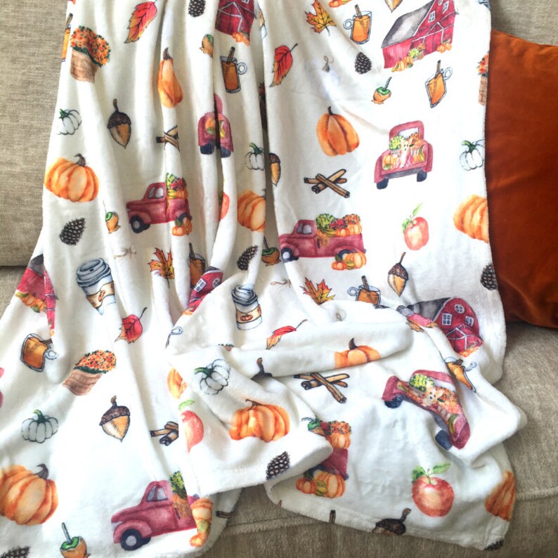May include: A white throw blanket featuring a repeating watercolor pattern of red pickup trucks carrying pumpkins, red barns, orange pumpkins, acorns, pinecones, cinnamon sticks, apples, mugs of cider, and potted orange flowers.
