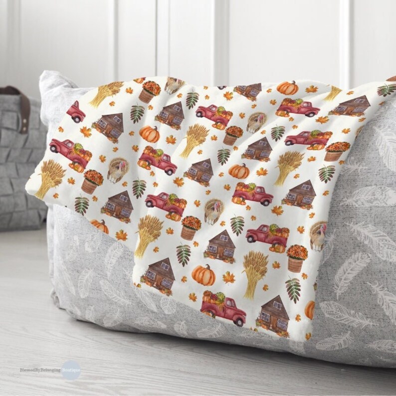 May include: A white throw blanket featuring a repeating watercolor pattern of red pickup trucks carrying pumpkins, brown wooden cabins, golden wheat sheaves, orange pumpkins, potted autumn flowers, turkeys, and scattered colorful fall leaves.