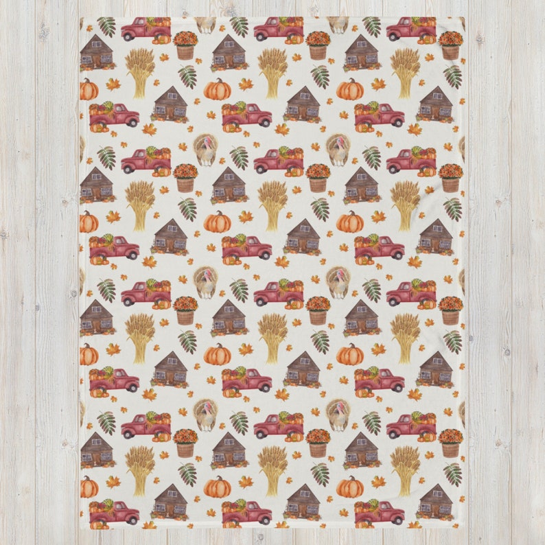 May include: A cream-colored throw blanket featuring a repeating autumn-themed pattern. The design includes illustrations of red pickup trucks carrying pumpkins, brown wooden cabins, orange pumpkins, golden wheat sheaves, turkeys, and fall leaves.