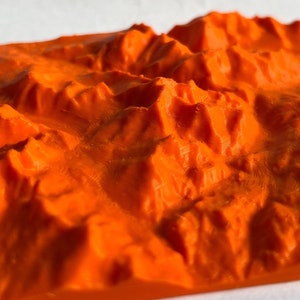 Puede incluir: Un modelo impreso en 3D de una cordillera, renderizado en un color naranja brillante. El modelo está detallado con crestas y picos, creando una representación realista de un paisaje montañoso.