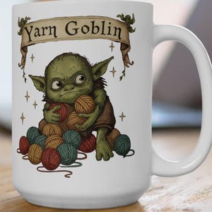 Peut inclure: Mug en c&eacute;ramique blanche avec une illustration de gobelin vert tenant des pelotes de laine. Le mug porte l'inscription "Yarn Goblin" sur une banni&egrave;re. Le gobelin est entour&eacute; de pelotes de laine rouges, turquoise et marron.