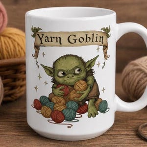 Peut inclure: Mug en c&eacute;ramique blanche avec un personnage de dessin anim&eacute; vert tenant des pelotes de laine. Le mug affiche le texte "Yarn Goblin" sur une banni&egrave;re. Le design comprend des &eacute;toiles et diverses pelotes de laine color&eacute;es.