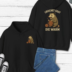 May include: Black hoodie with a front pocket and a graphic of an opossum knitting. The back of the hoodie features the text "CROCHET FAST DIE WARM" with an opossum knitting. A pair of blue jeans and black sneakers are also in the image.