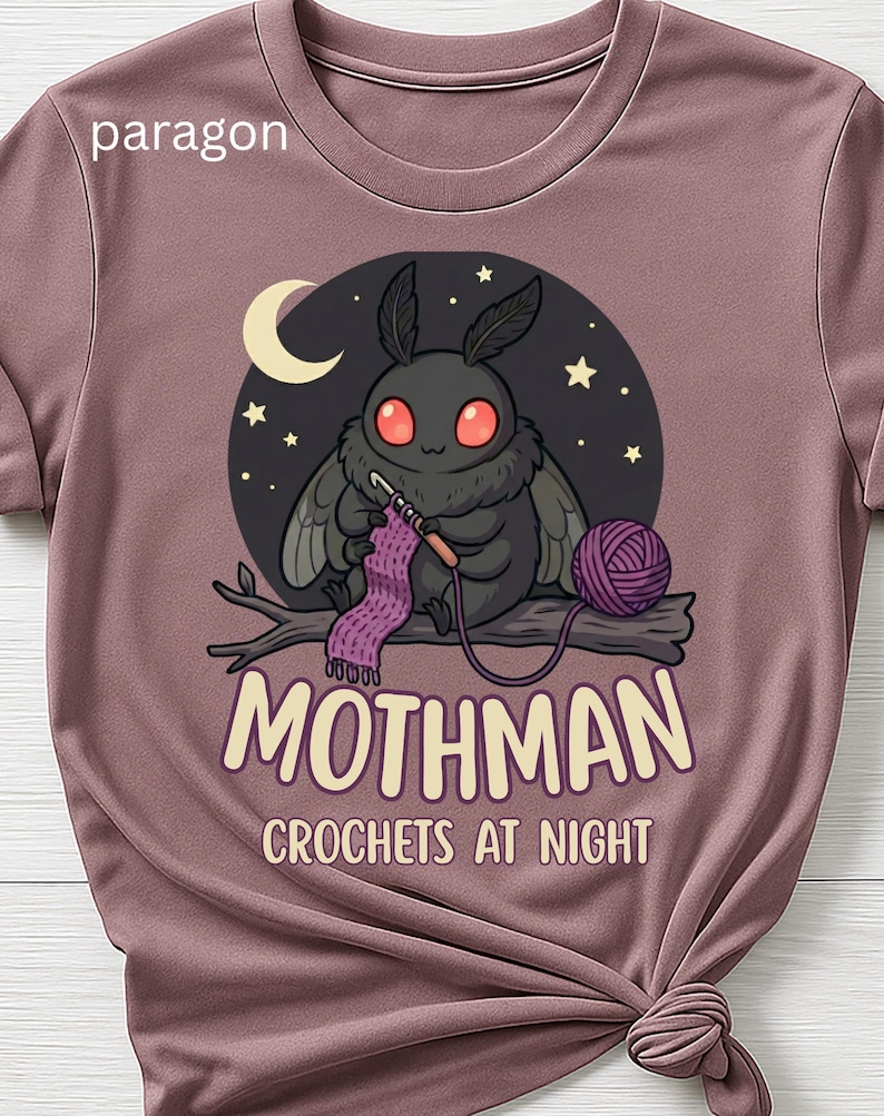 May include: Mauve t-shirt featuring a cartoon Mothman crocheting. The design includes a dark circle with a crescent moon, stars, and the text "Mothman Crochets at Night". A ball of yarn and a crochet project are also visible.