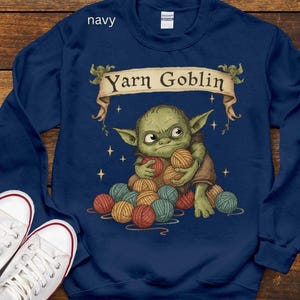 May include: Navy blue sweatshirt with a graphic of a green creature holding yarn balls. The text "Yarn Goblin" is displayed on a banner above the creature. White sneakers are visible in the lower left corner.
