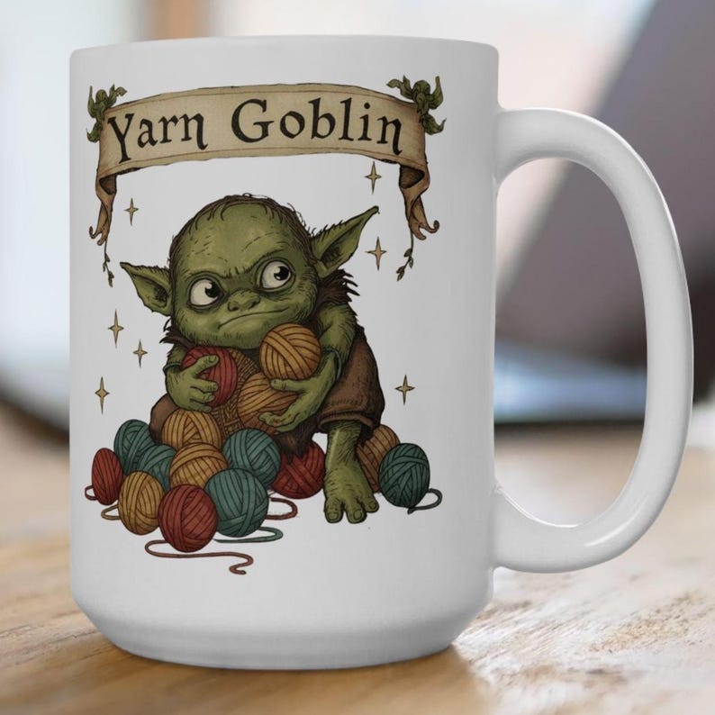 Peut inclure: Mug en c&eacute;ramique blanche avec une illustration de gobelin vert tenant des pelotes de laine. Le mug porte l'inscription "Yarn Goblin" sur une banni&egrave;re. Le gobelin est entour&eacute; de pelotes de laine rouges, turquoise et marron.